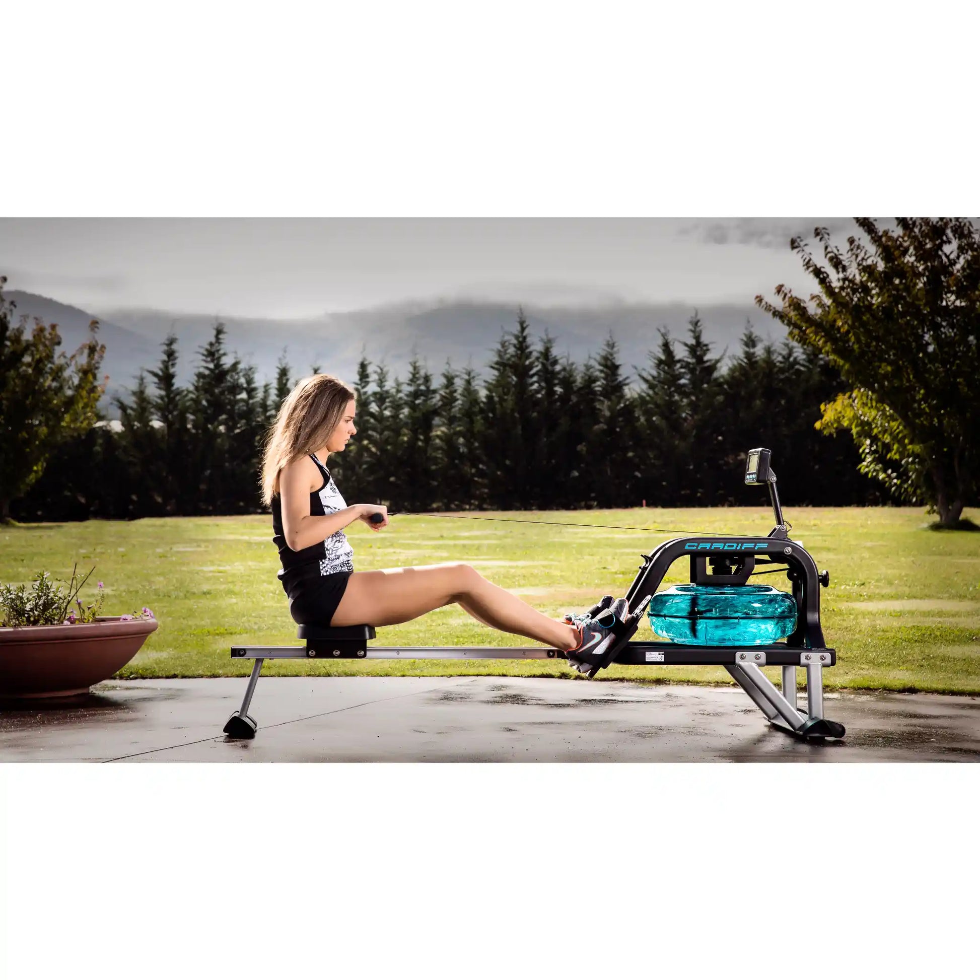 Woman exercising on a rowing machine outdoors with trees and mountains in the background