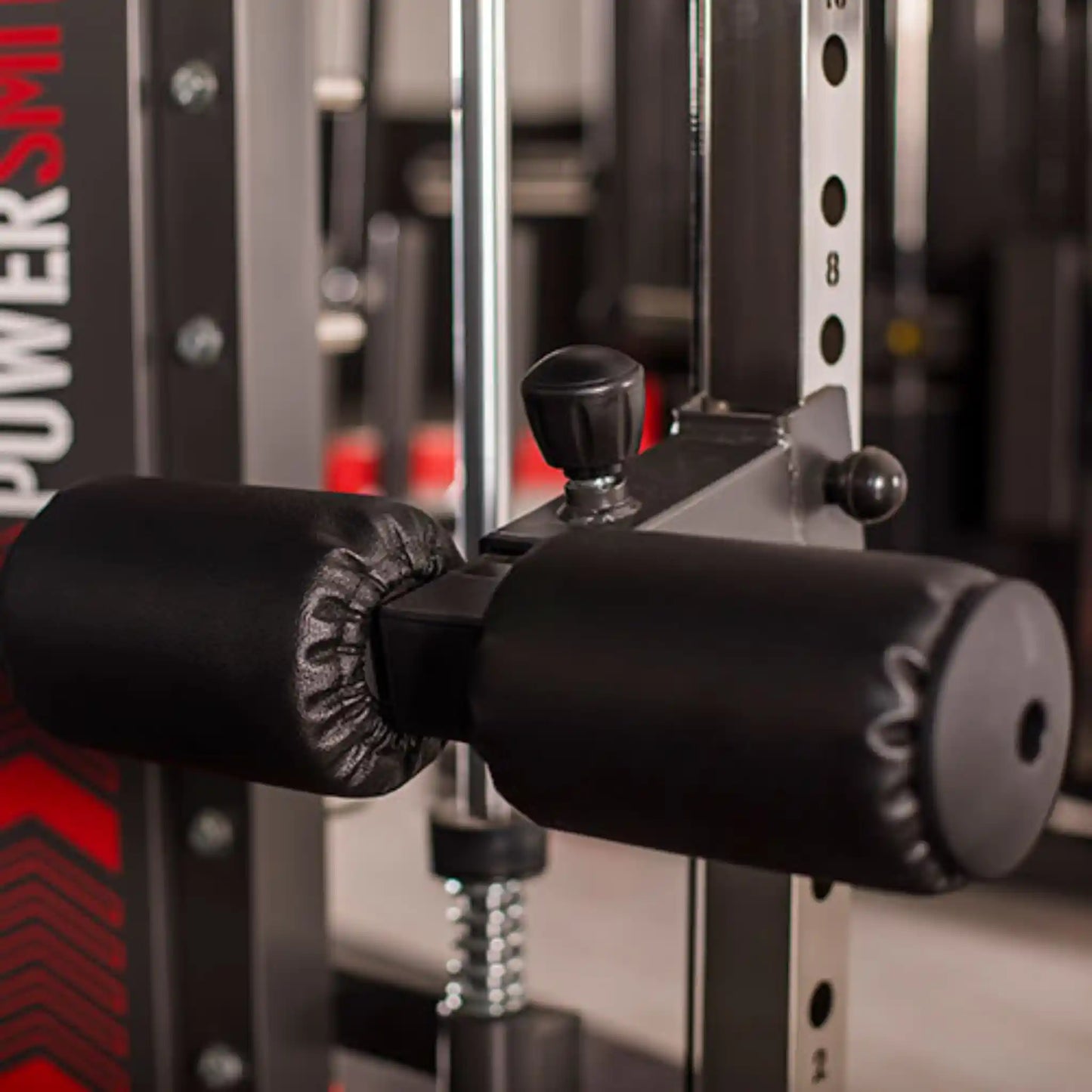 Close-up of a gym machine with black padding on handles, blurred gym equipment in the background.