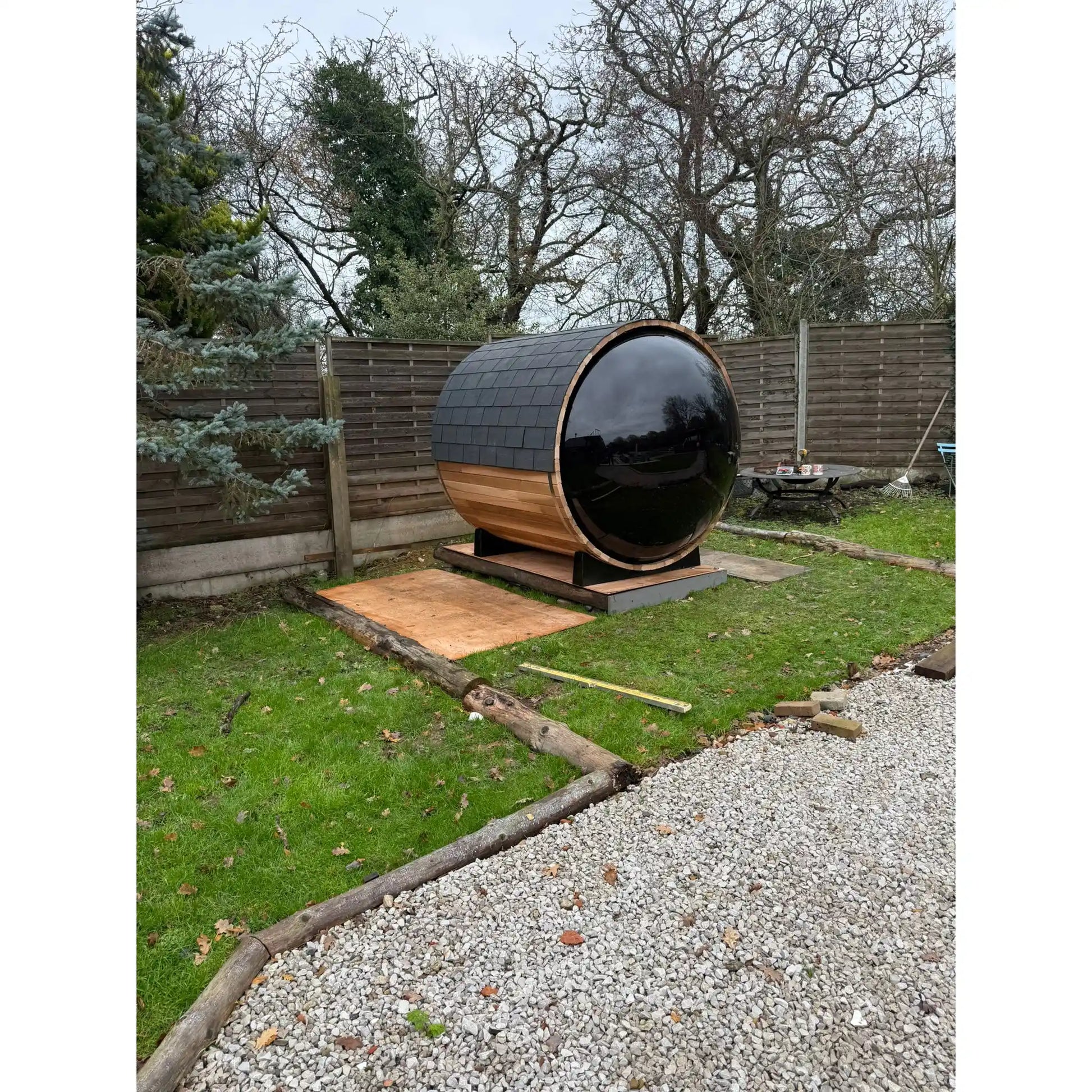 Large wooden barrel sauna with large black window in a garden setting with trees and a fence in the background.