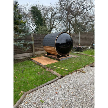 Large wooden barrel sauna with large black window in a garden setting with trees and a fence in the background.