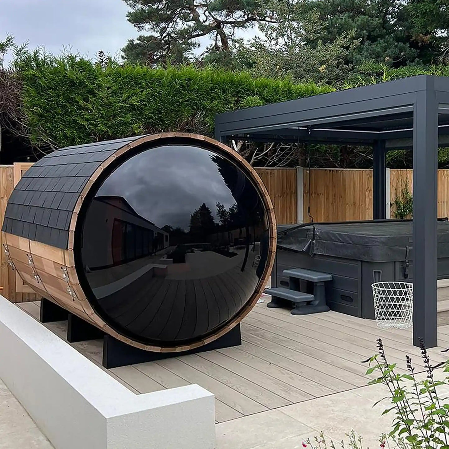 Wooden barrel sauna on a wooden deck with garden and trees in the background