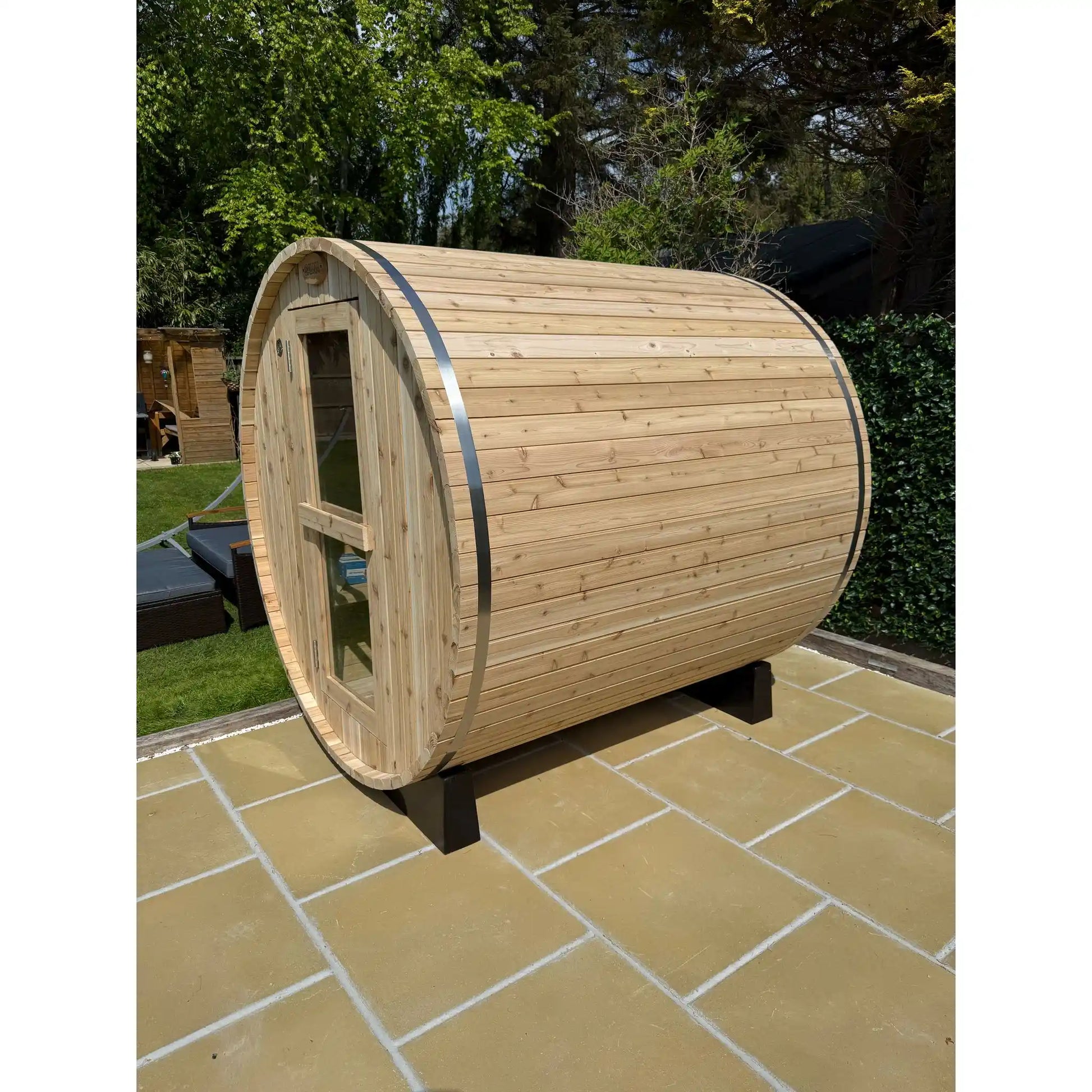 Wooden barrel-shaped sauna on a tiled patio with greenery in the background