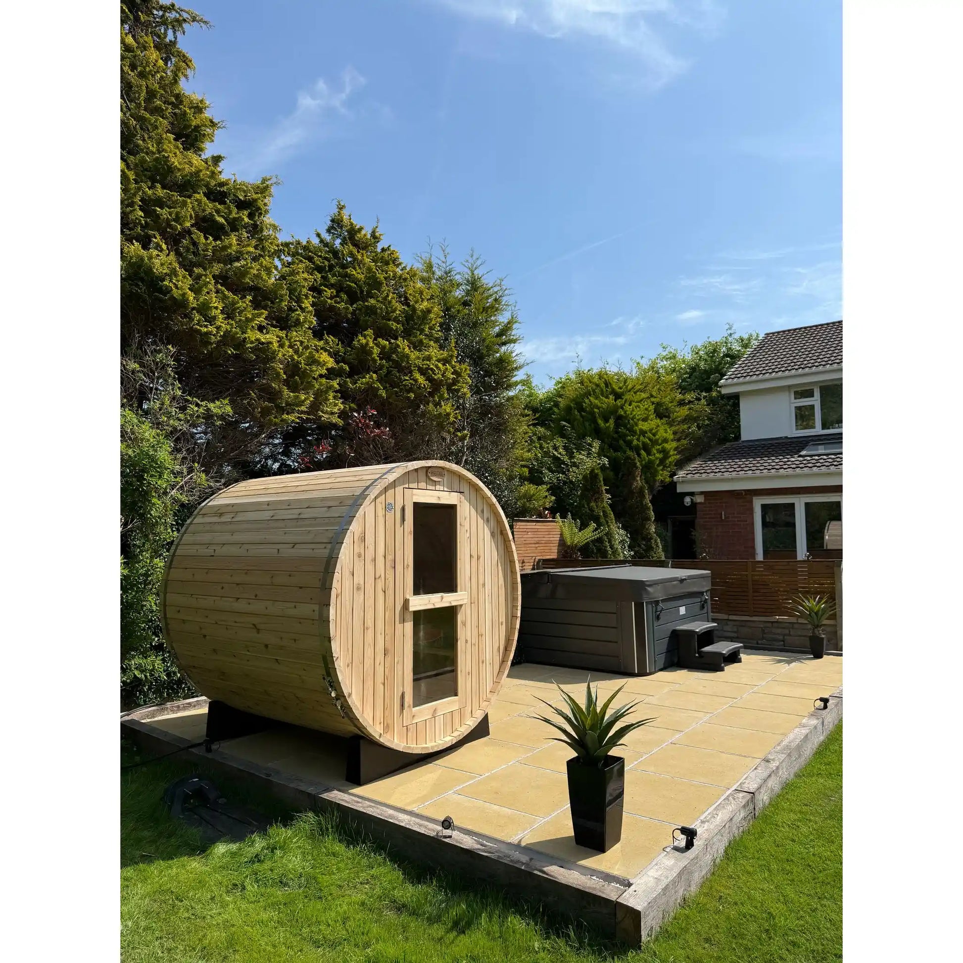Wooden barrel sauna in a garden setting with a hot tub and trees in the background.