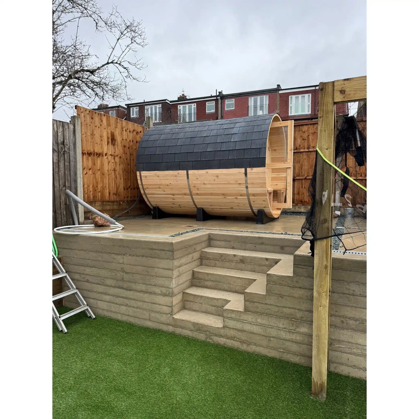 Wooden barrel sauna on a concrete platform with steps in a backyard setting.