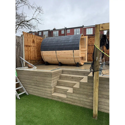 Wooden barrel sauna on a concrete platform with steps in a backyard setting.
