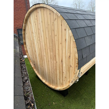 Back of a wooden barrel-shaped sauna with a roof on a grassy area.