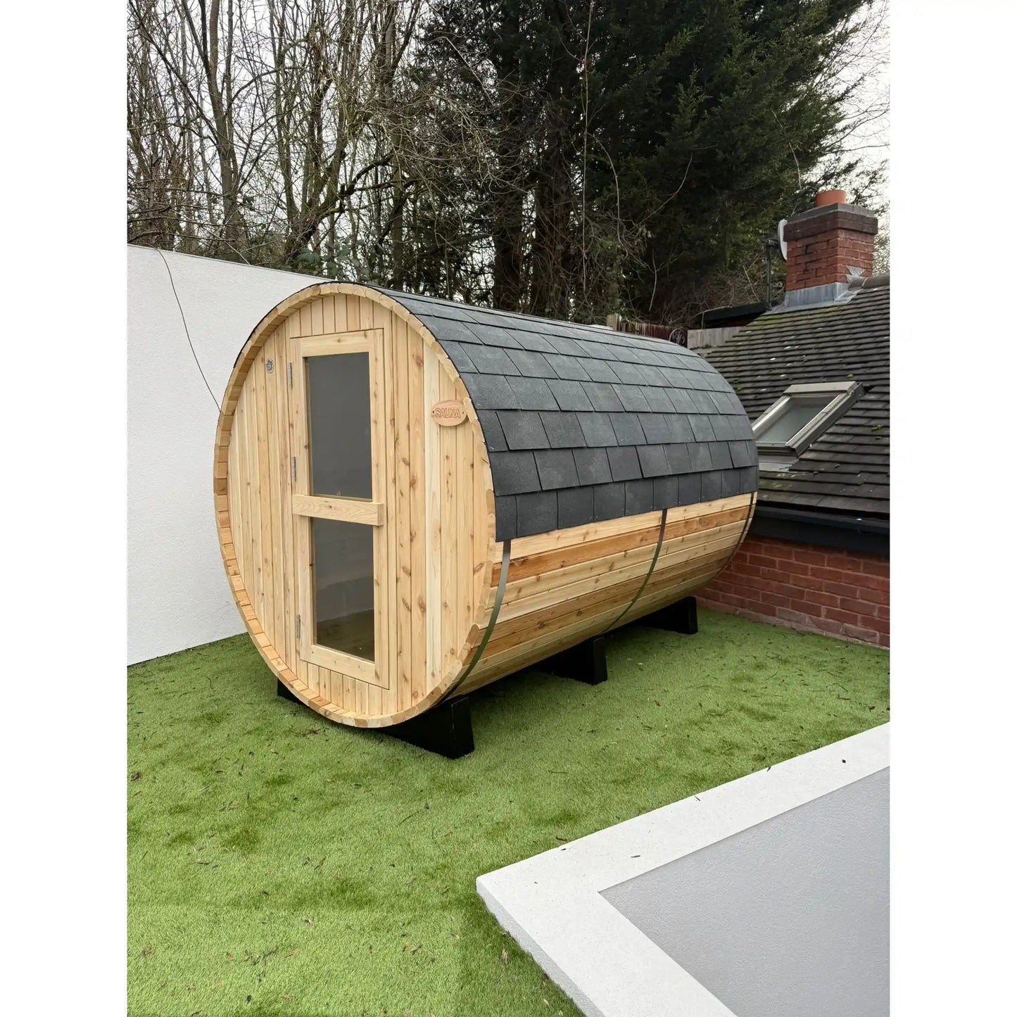 Wooden barrel-shaped sauna with a shingled roof on grass outside a house.