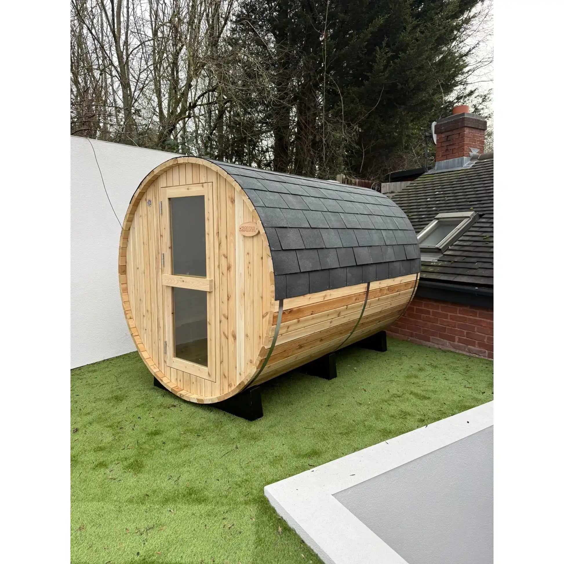 Wooden barrel-shaped sauna with a shingled roof on grass outside a house.