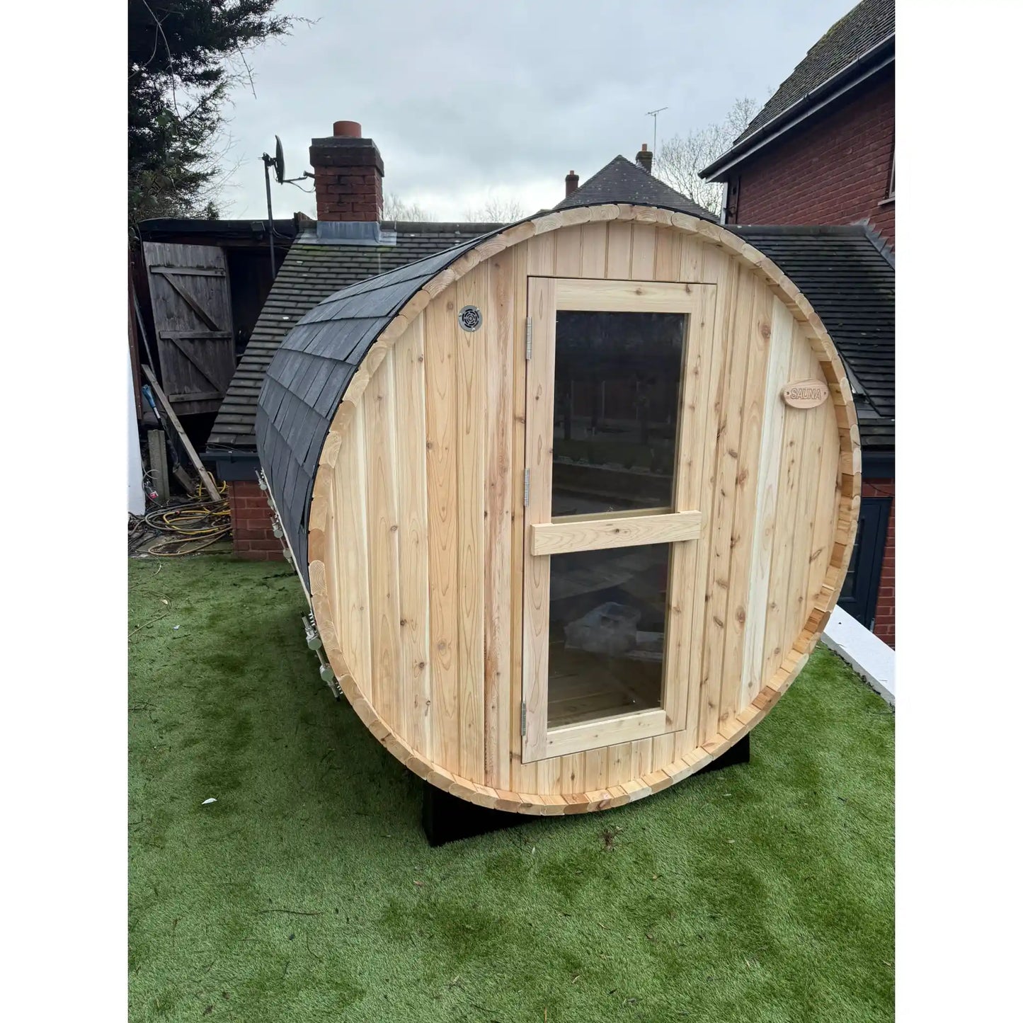 Wooden barrel-shaped structure on a grassy area with a house in the background