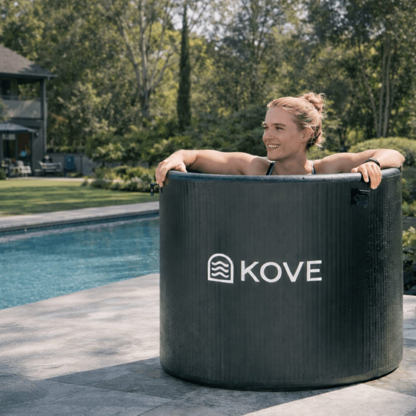 Woman sitting in an inflatable hot tub labeled 'Kove' by a pool with trees in the background