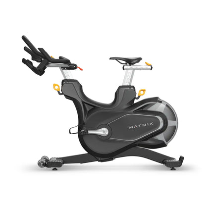 A Matrix brand indoor cycling machine in dark gray and black with silver metallic accents and small yellow details, shown from a three-quarters angle on a white background.