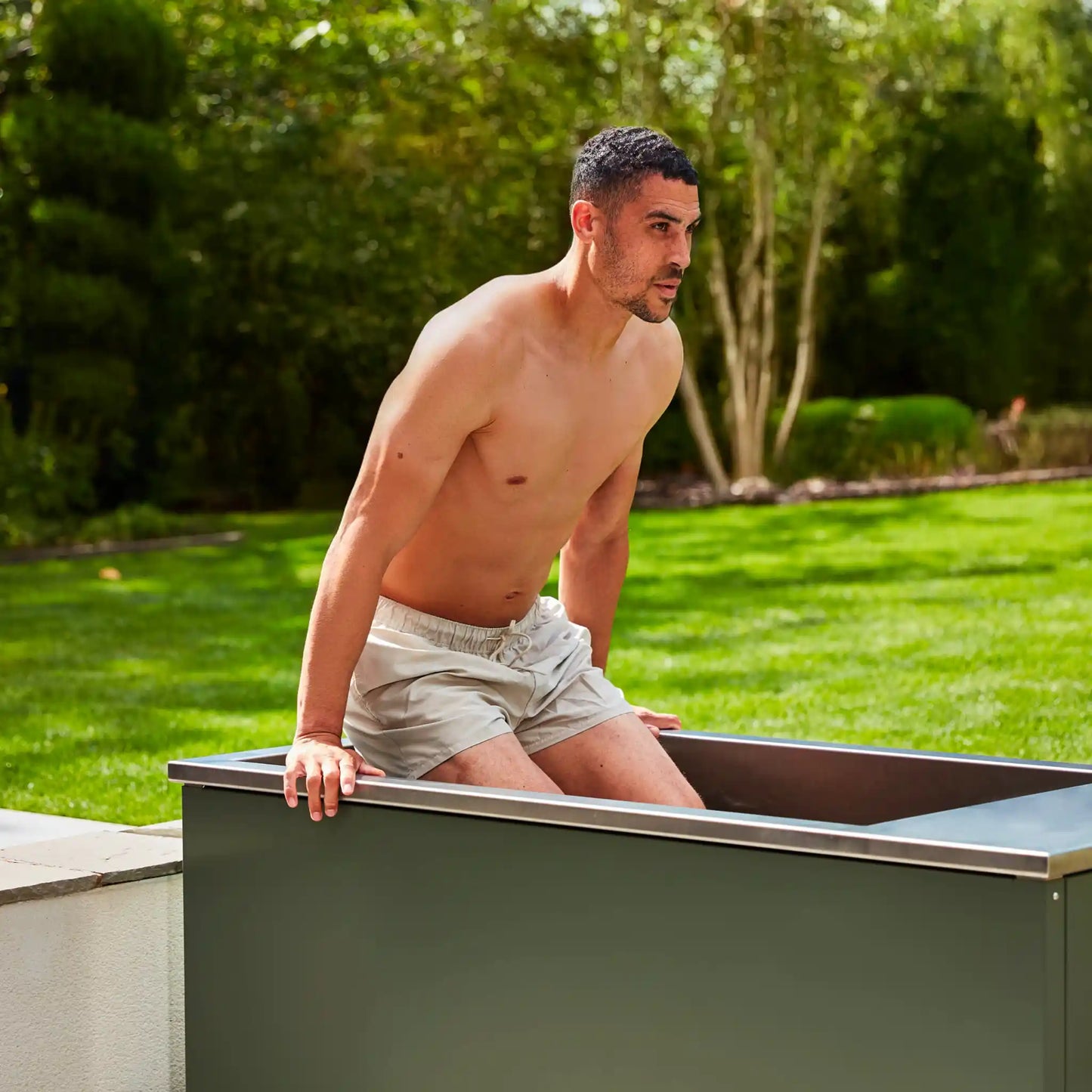 Man lowering himself into an ice bath outdoors with greenery in the background