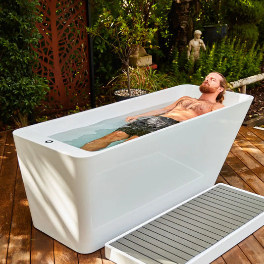 Man relaxing in a white ice tub on a wooden deck with greenery in the background