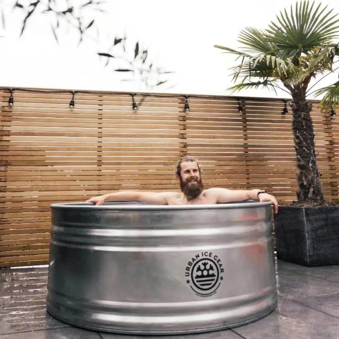 Man relaxing in a metal ice bath with 'Urban Ice Gear' logo outdoors.